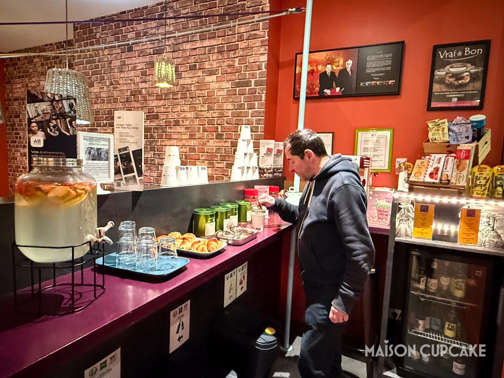 Hotel guest making himself herbal tea at self serve drinks counter in Ibis Styles Lille Grand Place hotel's colourful lobby.