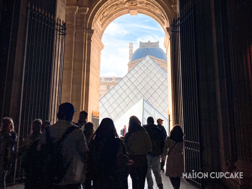 Glass pyramid and crowds at the Louvre, seen through archway.