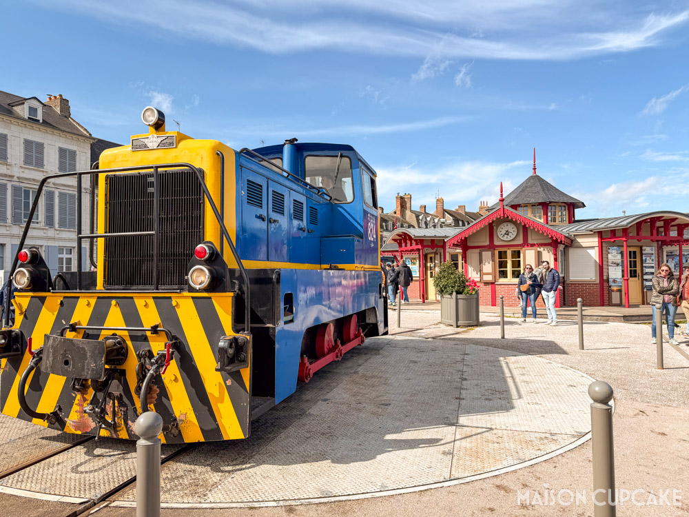 Bright blue and yellow locomotive of heritage steam railway at Saint-Valery-sur-Somme in northern France, with red station ticket booth behind on a clear sunny day.