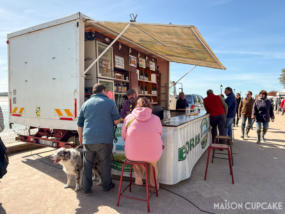 Harbourside beer stall with two customers and dog talking to owner, at Le Crotoy, northern France - the sky is clear blue. 