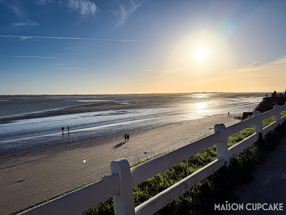 Sunset at Le Crotoy with sunlight reflecting on water as figures in distance walk on the sand, with white fence in foreground